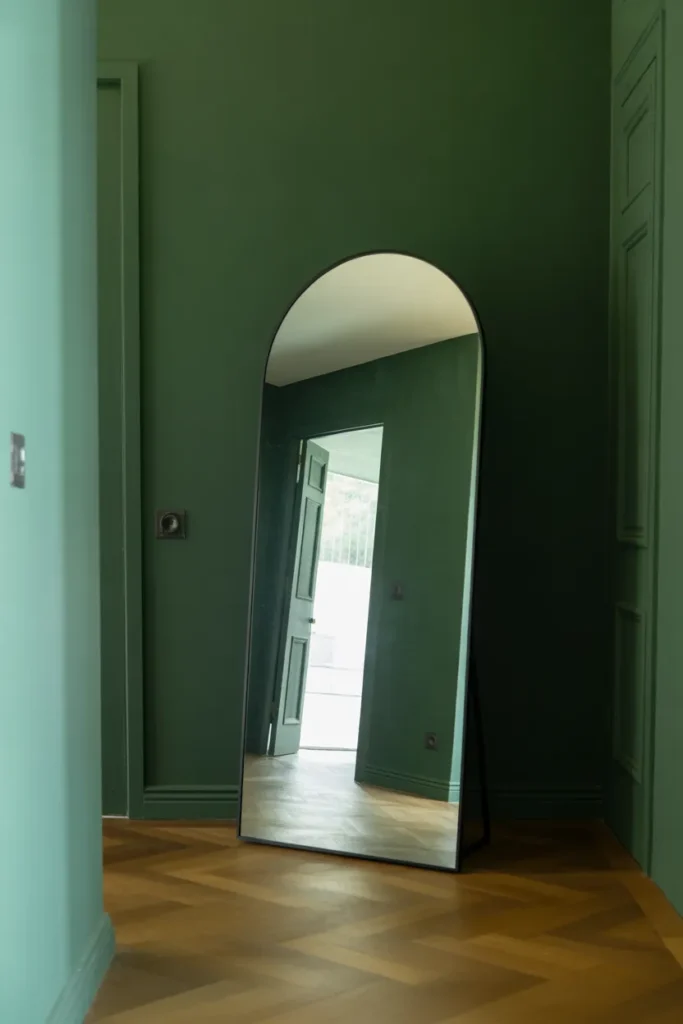 A tall arched floor mirror leaning against a deep sage green wall in a foyer