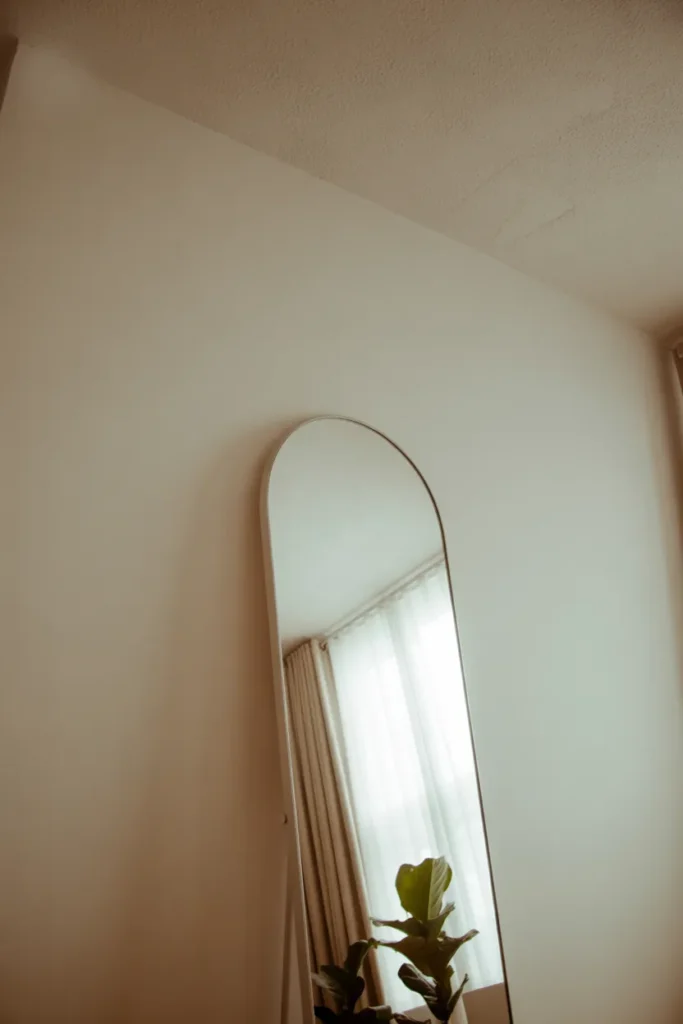 A tall arched mirror leans against a warm white wall beside a curtained window